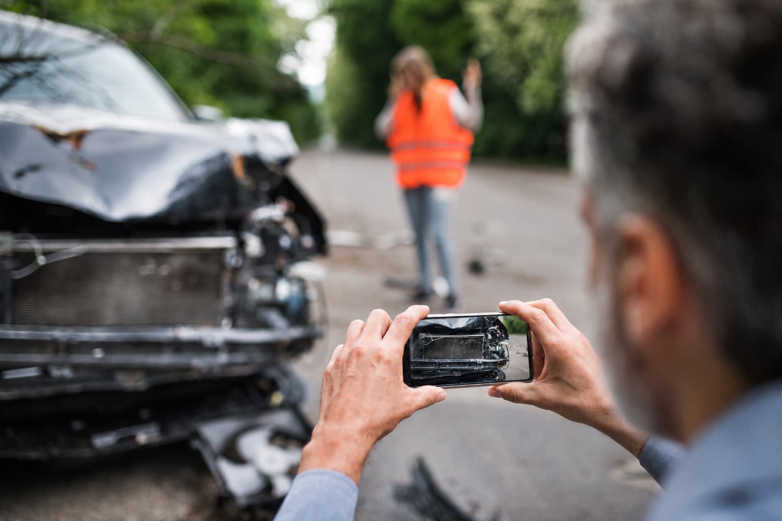 A man photographing a badly damaged car on his phone, with another driver in a high visibility vest further up the road
