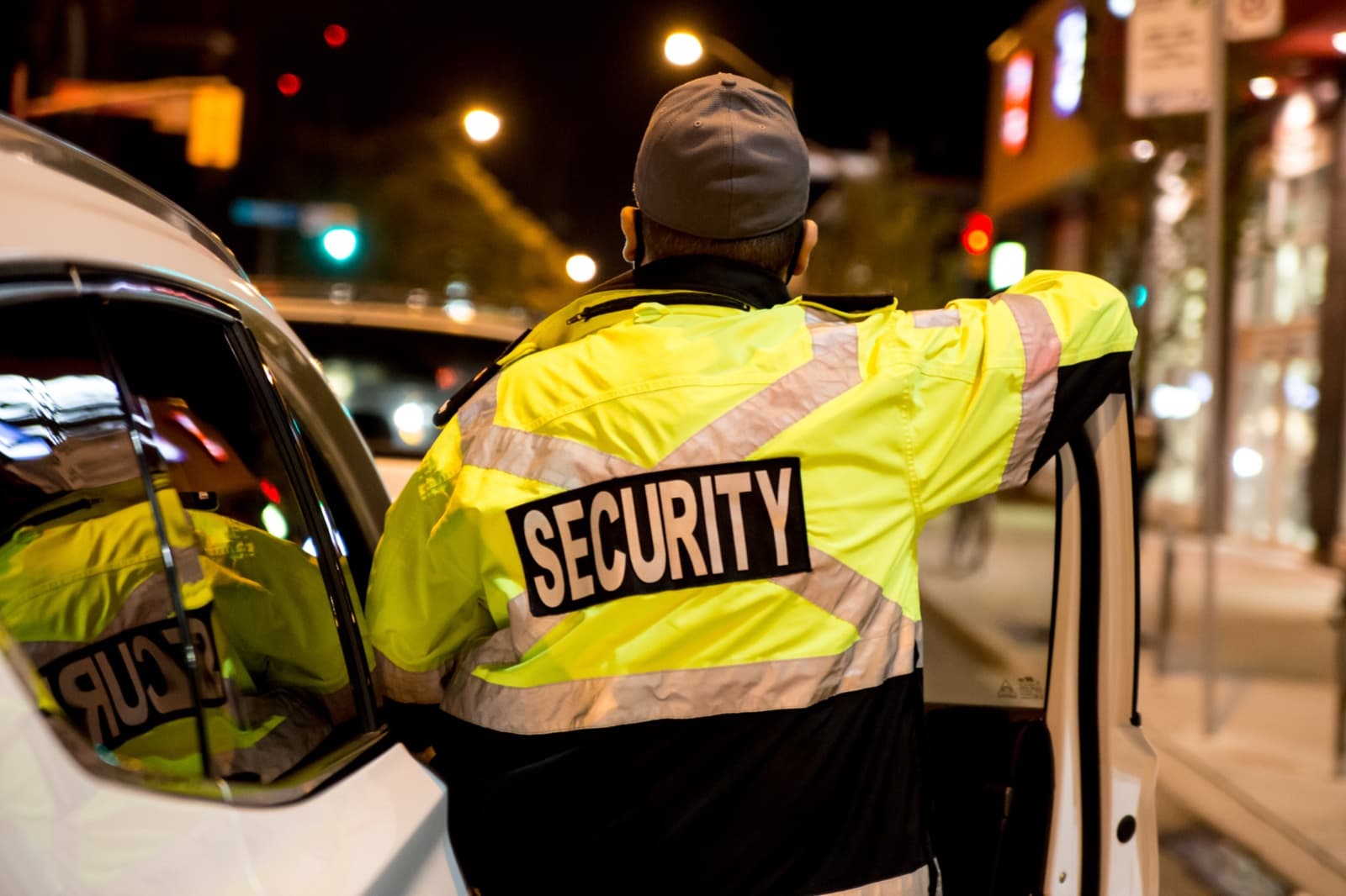A guard in a high visibility jacket marked Security, leaning on a vehicle door on a street at night