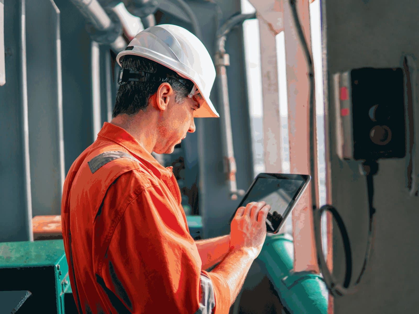 An inspector in a hard hat and orange coveralls records an inspection on a tablet beside plant pipework.