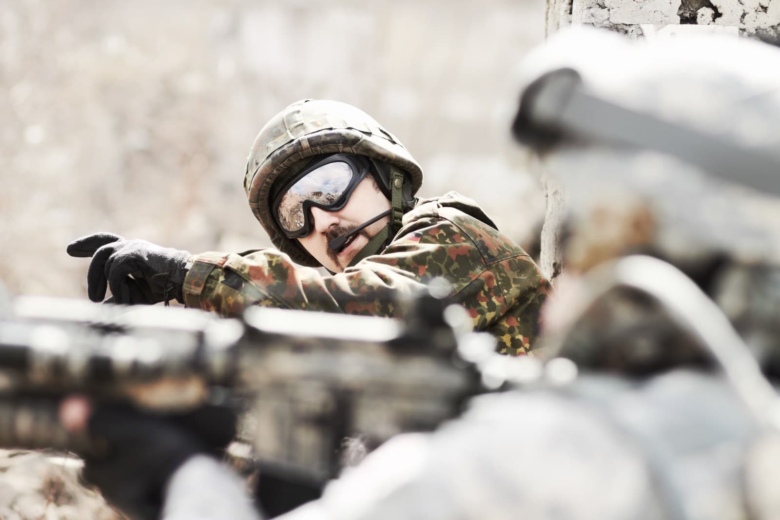 A soldier in helmet and goggles signalling from cover, a rifle in the foreground.