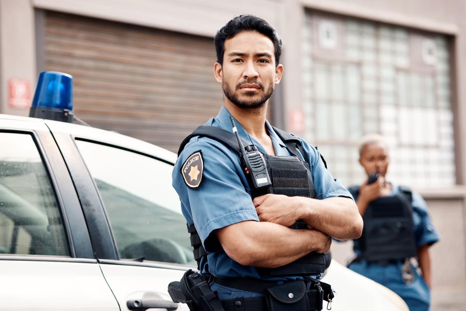 A police officer standing beside a marked car, vest and radio worn, a second officer behind.
